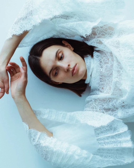 Young woman with brown hair lying down in a white lace dress, close-up shot, minimal background. Soft, diffused lighting creates a serene mood.