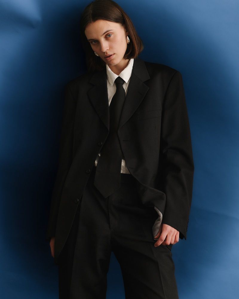 Woman wearing an oversized black tailored suit, white shirt, and black tie against a vibrant blue studio backdrop.