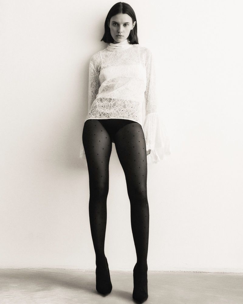 A slender adult woman in black polka-dot tights and a white lace top poses against a white wall in a studio environment.