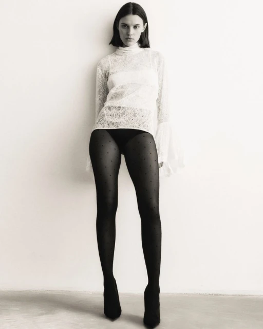 A slender adult woman in black polka-dot tights and a white lace top poses against a white wall in a studio environment.