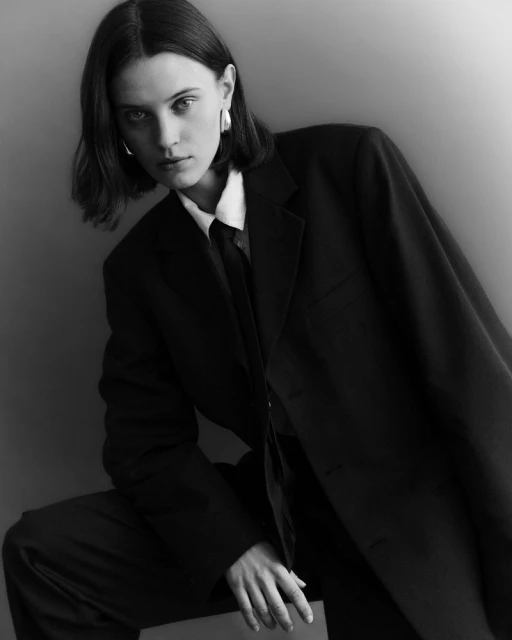 Black and white portrait of a young woman in a dark suit, posed indoors with silver earrings, looking at the camera in studio lighting.