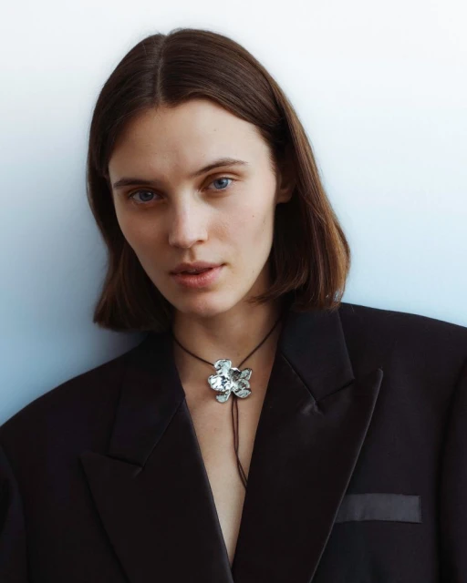 Portrait of a woman with shoulder-length brown hair wearing a black blazer and a large silver flower necklace against a light blue background.