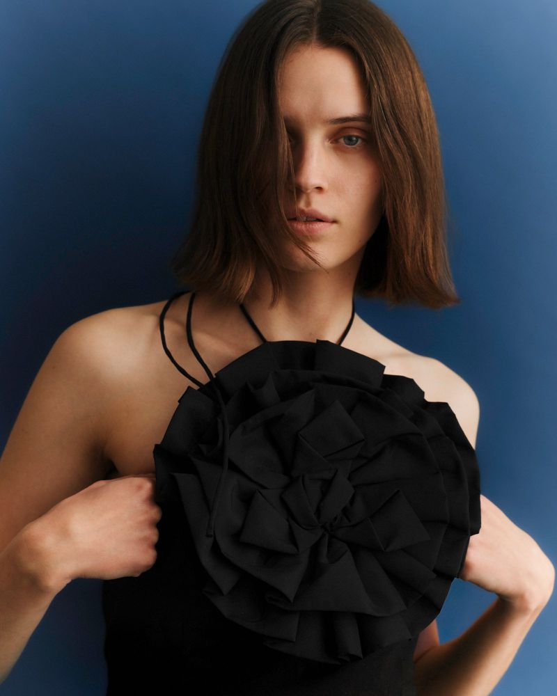 Portrait of a woman with short brown hair wearing a black top with a large, ruffled floral detail against a dark blue backdrop.