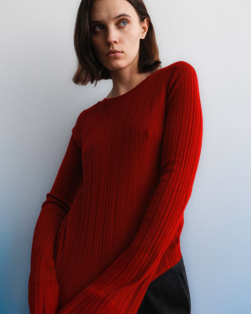 Portrait of a young woman with short brown hair wearing a red, long-sleeved, ribbed knit shirt, posing in front of a graduated blue wall.