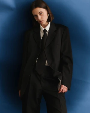 Woman wearing an oversized black tailored suit, white shirt, and black tie against a vibrant blue studio backdrop.