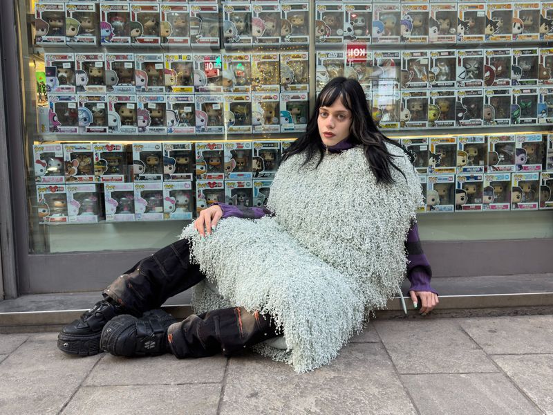 Woman in a fuzzy garment poses outdoors in front of window display with rows of Funko Pop boxes, seated on a gray city sidewalk.