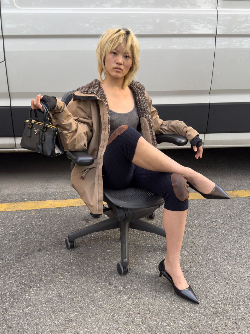 A woman sits on an office chair in a parking lot, wearing a jacket and leggings, and holding a handbag with a white van behind her.