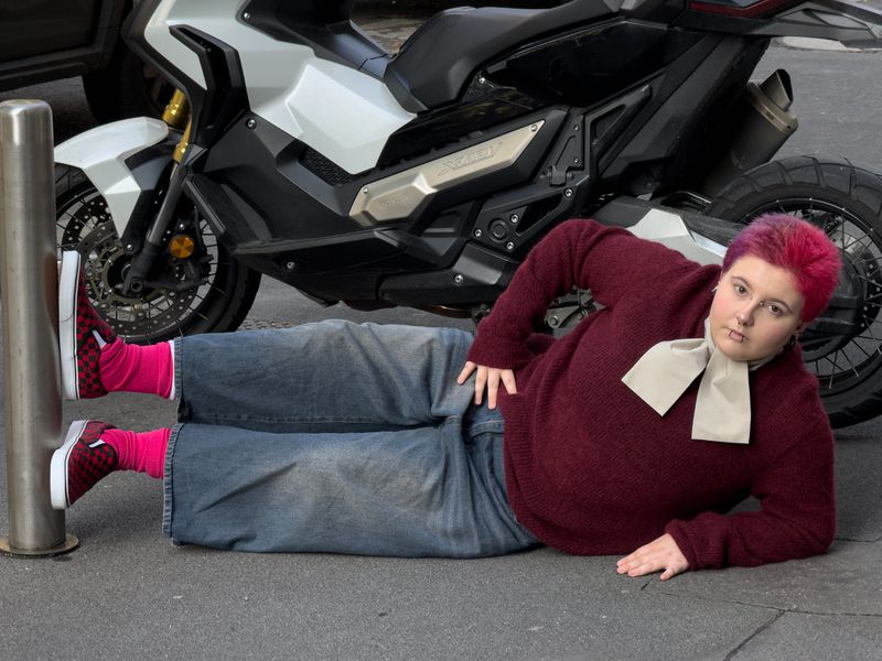 Person poses on pavement with a motorcycle in the background, wearing jeans, a maroon sweater, colorful socks, and red shoes.