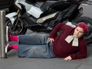 Person with pink hair lying on the street in a burgundy sweater and wide-leg jeans, propping feet on a metal bollard.