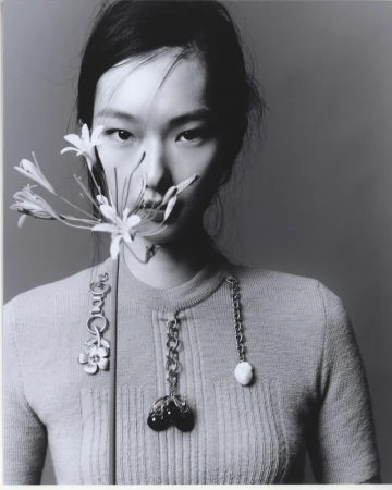 Black and white portrait of a woman wearing a knitted top, holding white flowers near her face, with decorative chain pendants.