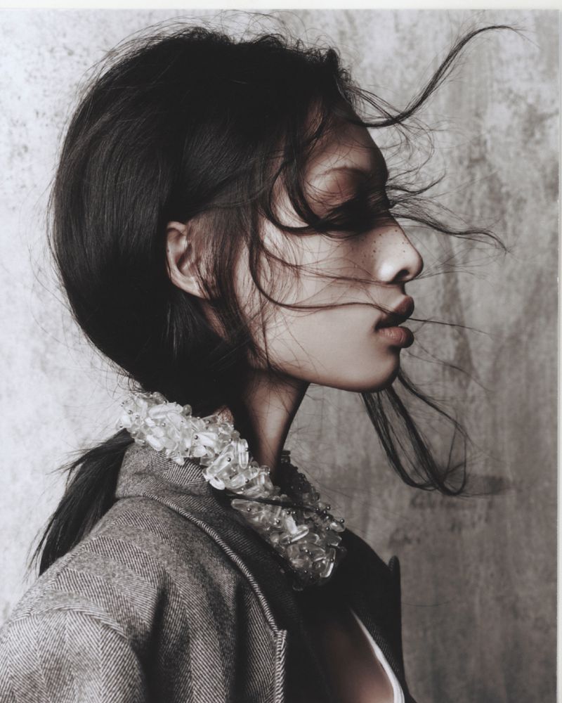 Profile portrait of an adult woman with dark hair in a wool coat with a beaded collar against a textured grey background.