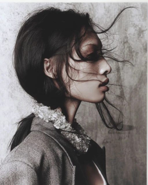 Profile portrait of an adult woman with dark hair in a wool coat with a beaded collar against a textured grey background.