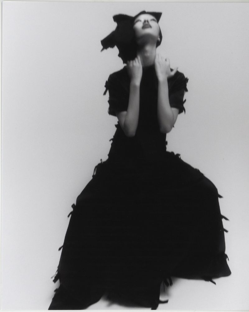 Woman in a black dress with her hands around her neck, looking upwards, against a plain white background in a studio shot.