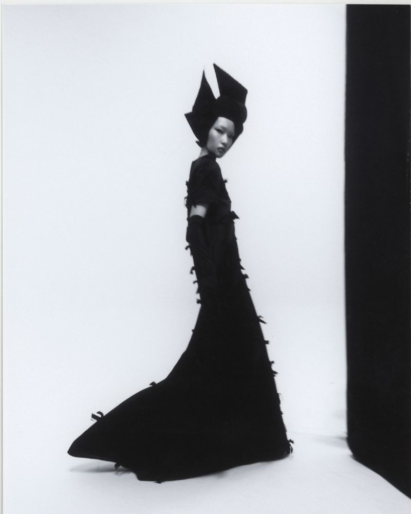 A striking black-and-white fashion shot of an Asian woman in a black couture gown and avant-garde headpiece, posing against a graduated background.