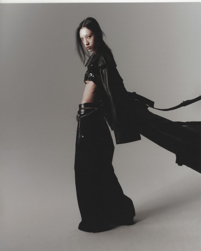 A slender person with dark hair poses in a dark, flowing outfit against a neutral backdrop, captured with dramatic flair in a studio setting.