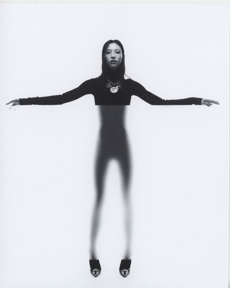 Model with outstretched arms in a black long-sleeve top and statement necklace against a white seamless backdrop.