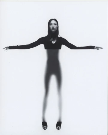 Model with outstretched arms in a black long-sleeve top and statement necklace against a white seamless backdrop.
