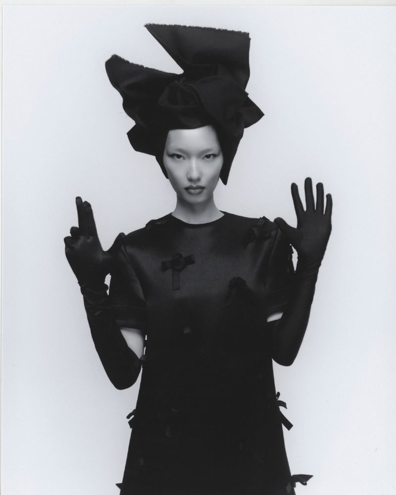 Woman in oversized black bow headpiece and satin dress striking a dramatic pose against a stark white backdrop in a fashion shot.
