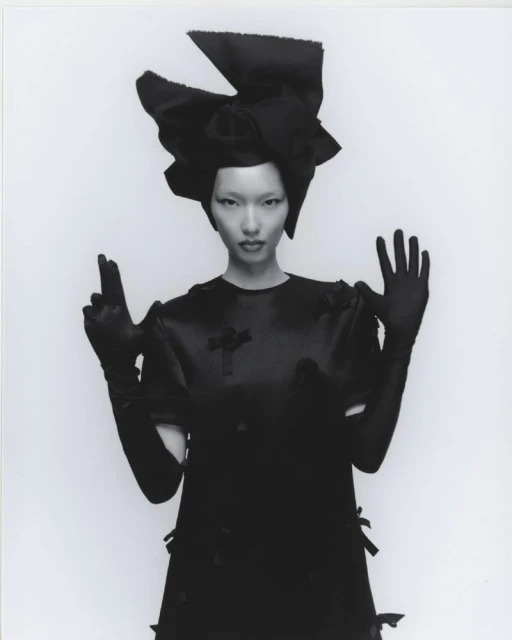 Woman in oversized black bow headpiece and satin dress striking a dramatic pose against a stark white backdrop in a fashion shot.