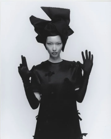 Woman in oversized black bow headpiece and satin dress striking a dramatic pose against a stark white backdrop in a fashion shot.
