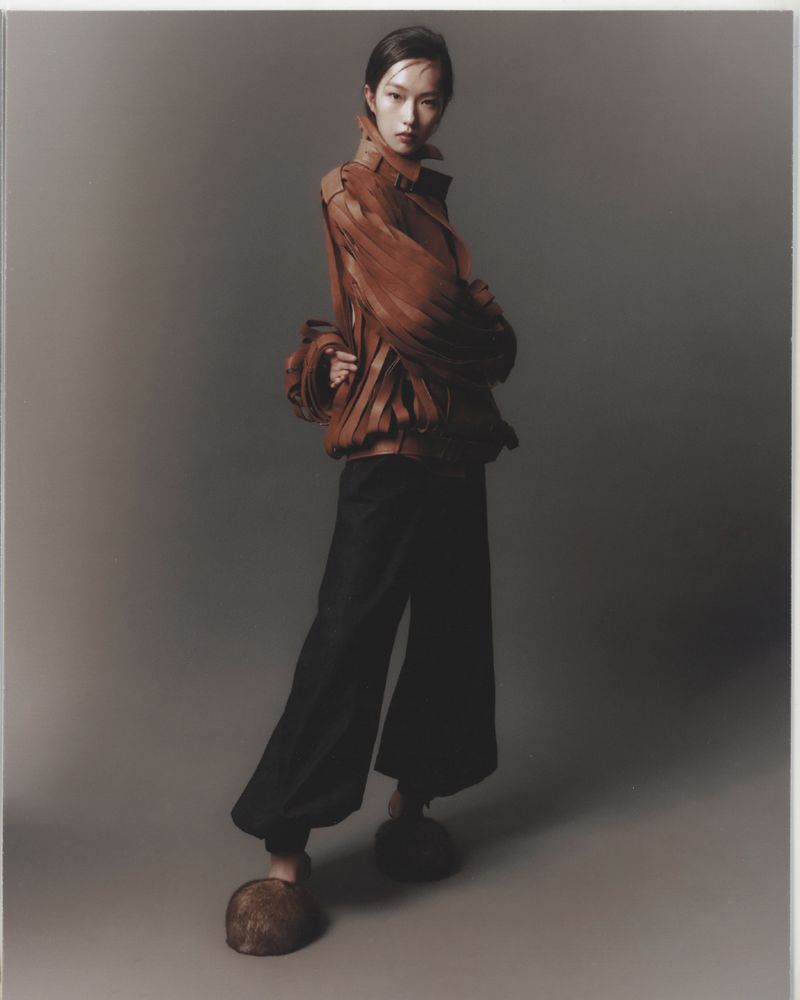 Studio shot of an Asian woman in a brown leather jacket, black pants, and wooden platform shoes posing in a fashion forward style.
