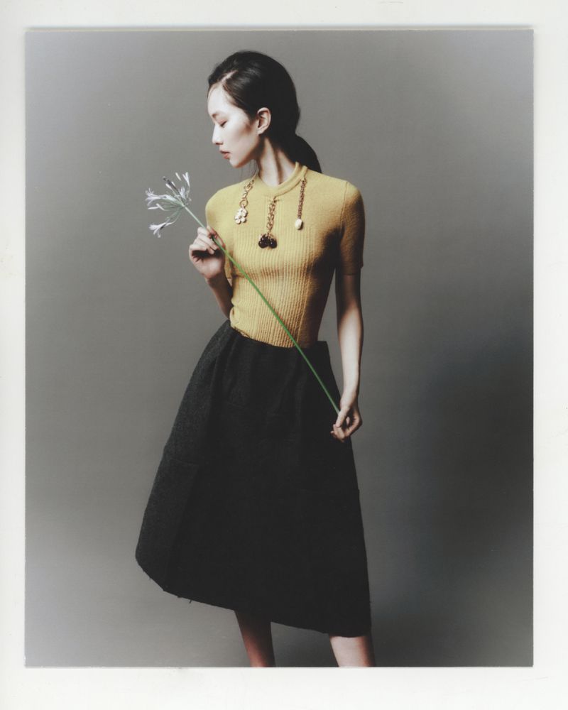 Asian woman in yellow sweater and black skirt holding a flower in a studio with gray backdrop, wearing gold necklace, fashion editorial.
