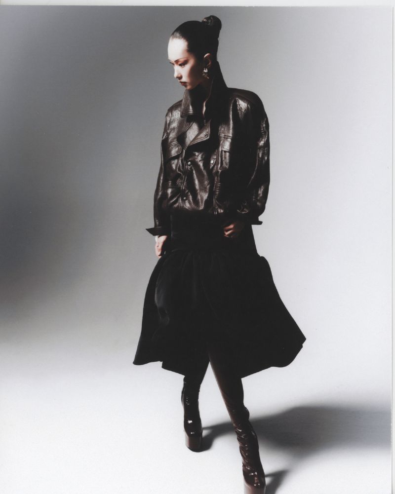 Fashionable woman in a brown leather jacket, black pants, and high heel boots poses in a photo studio with a neutral gray backdrop.