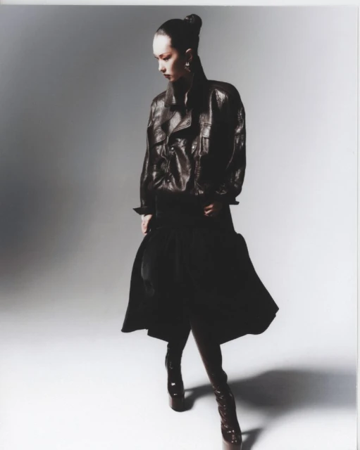Fashionable woman in a brown leather jacket, black pants, and high heel boots poses in a photo studio with a neutral gray backdrop.