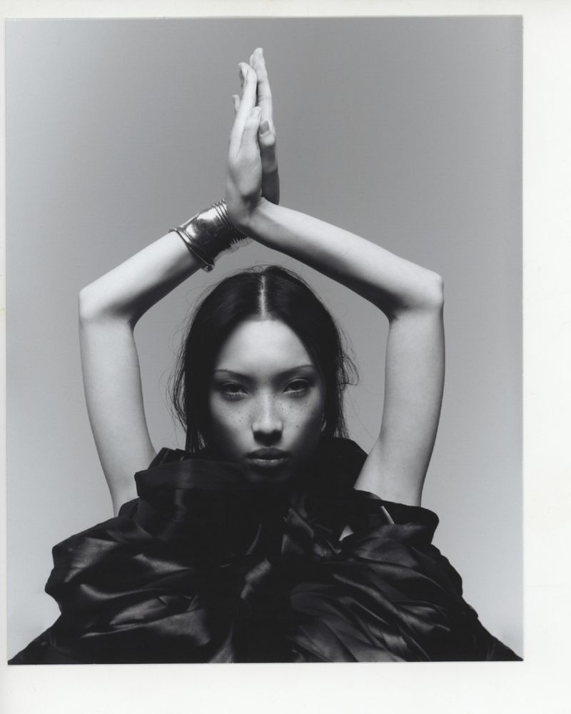 Black and white portrait of a woman in black fabric, hands above head, silver bracelet, against a grey backdrop in a photo studio.