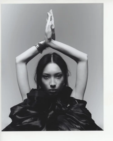 Black and white portrait of a woman in black fabric, hands above head, silver bracelet, against a grey backdrop in a photo studio.