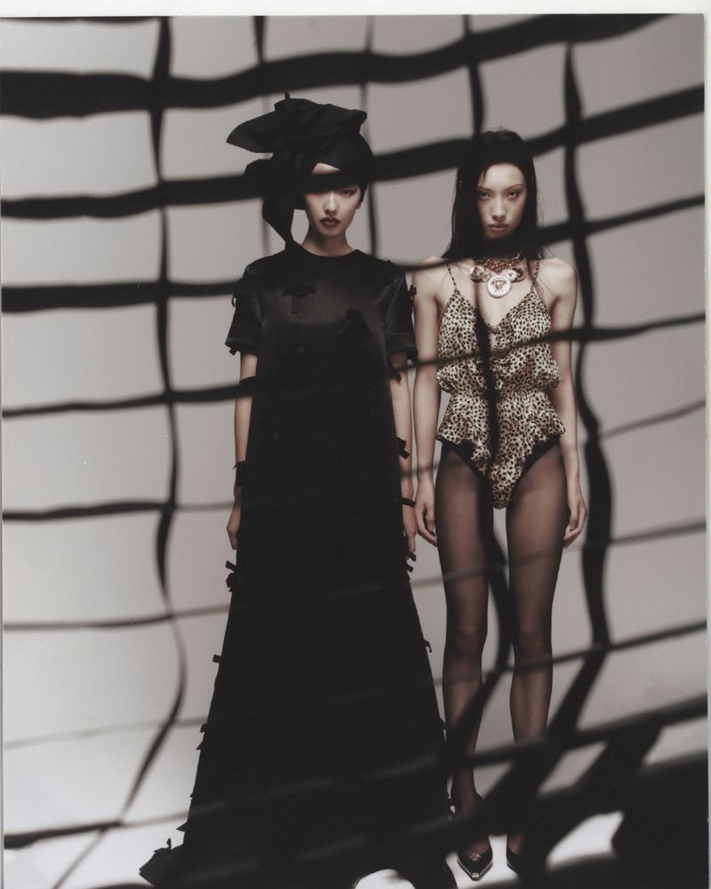 Two Asian fashion models pose in a studio wearing contrasting dresses: one in a long black dress, and the other in a leopard print swimsuit.
