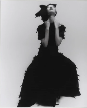 Model in a black textured gown and dramatic headpiece looking upward against a pale studio backdrop.