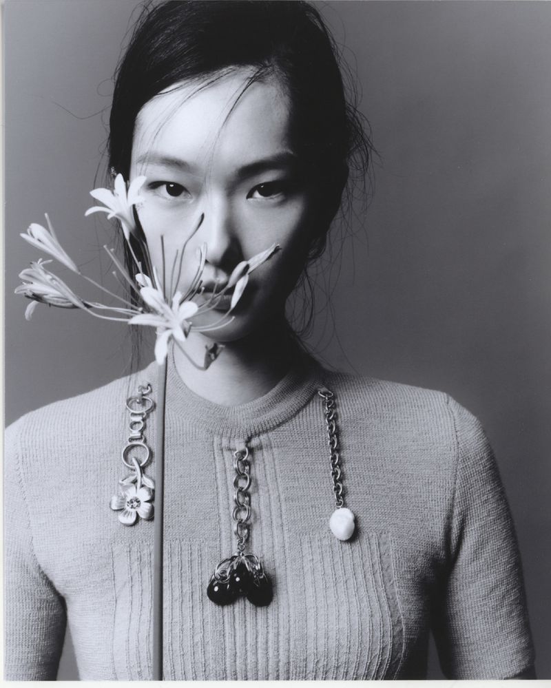 Black and white portrait of a woman wearing a knitted top, holding white flowers near her face, with decorative chain pendants.
