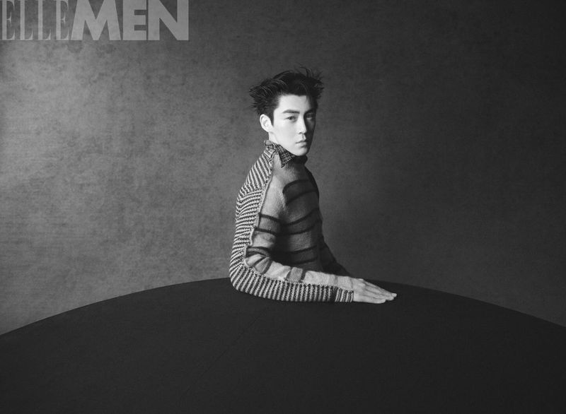 Black and white portrait of a person with a patterned top sitting at a table, set against a solid backdrop, capturing a contemplative mood.