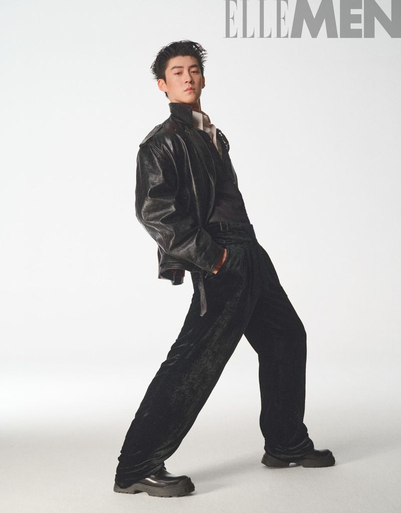 A stylish Asian male model in a black leather jacket and velvet pants poses confidently in a fashion studio against a white background.