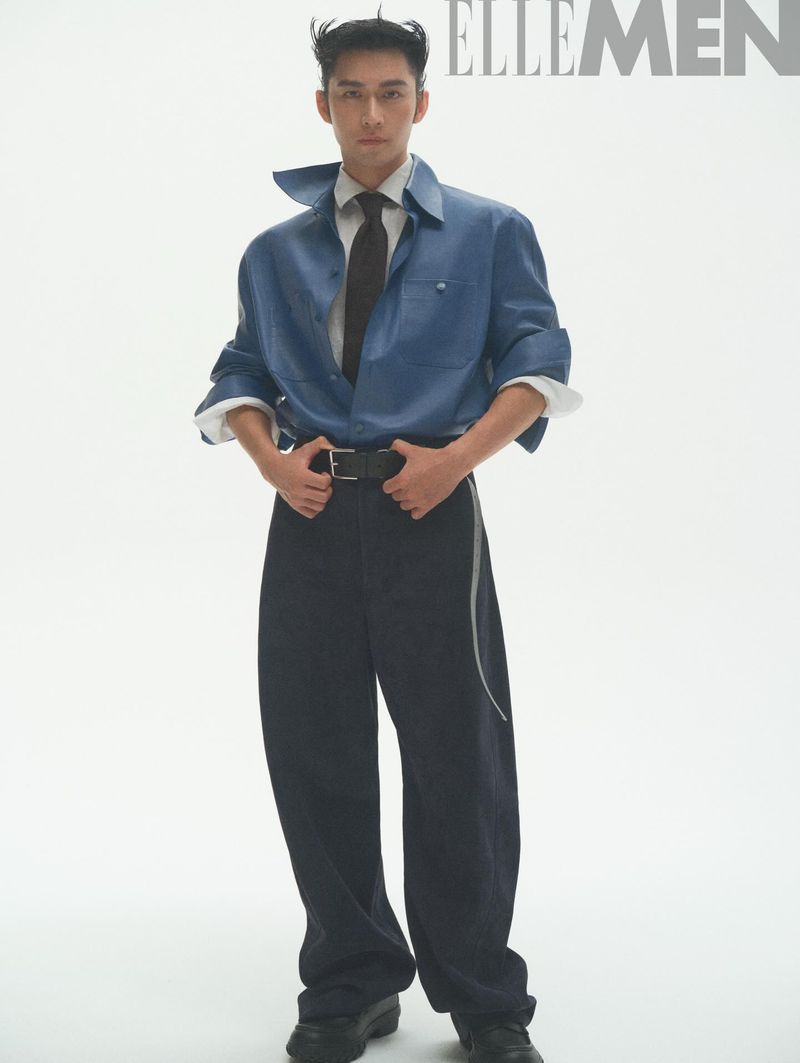 East Asian man standing in a blue collared shirt, dark tie, and baggy black trousers against a white studio background.