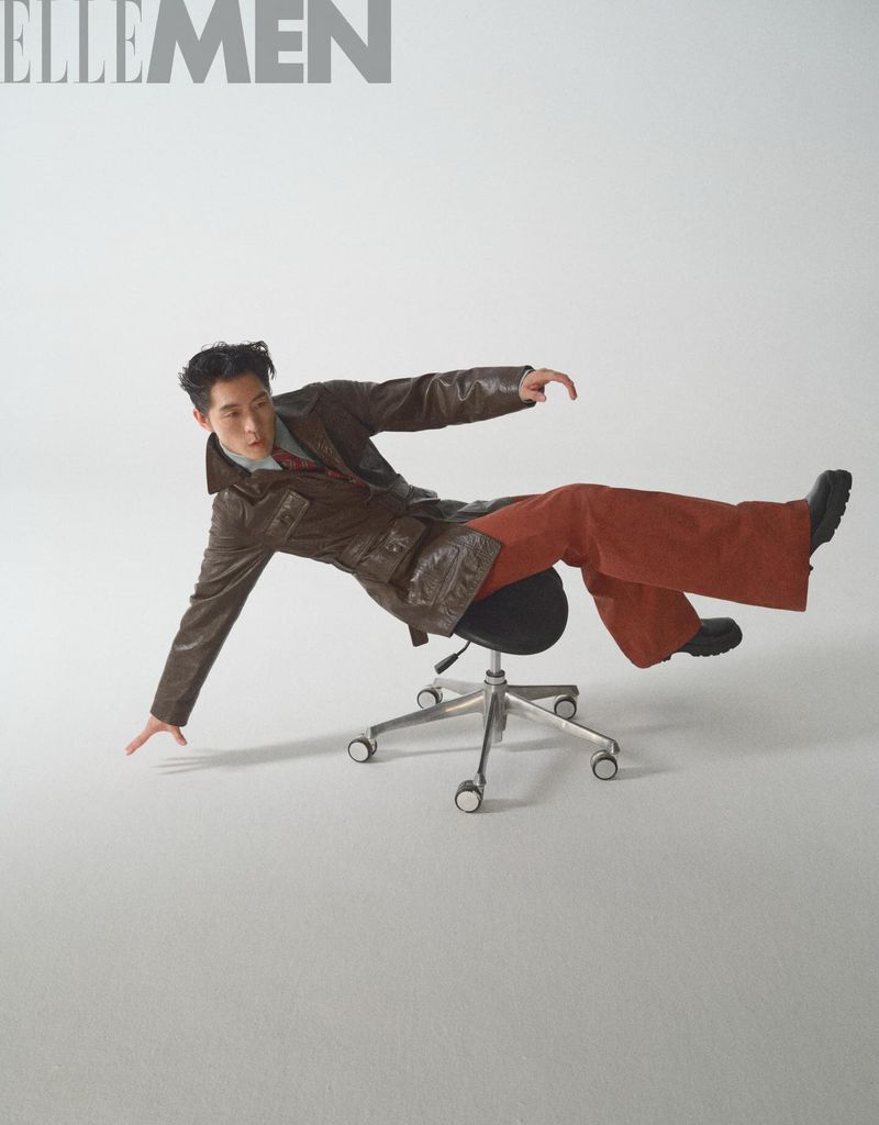 Adult male poses on a rolling chair in a studio setting. He wears a brown jacket and rust-colored pants for an editorial shot.