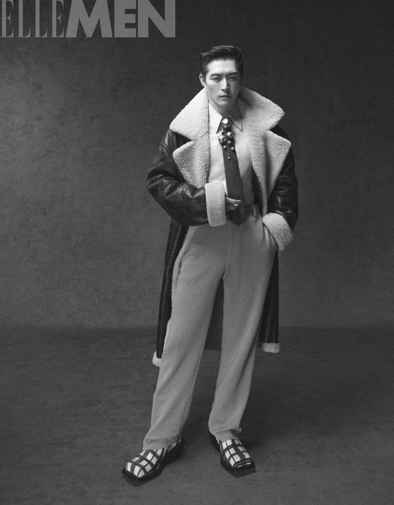 Monochrome portrait of a stylish adult male in a fur-lined coat, dress shirt, tie, and sandals, posing against a neutral background.