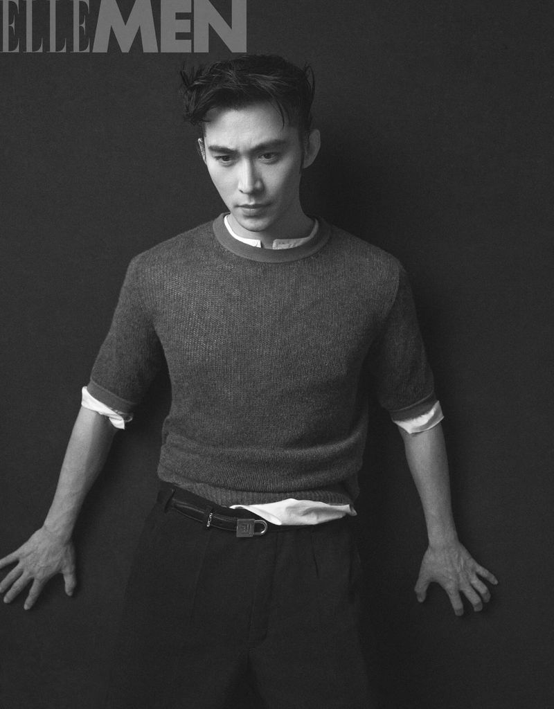 Black and white editorial portrait of an East Asian man wearing a short-sleeved knit sweater layered over a white shirt against a dark backdrop.