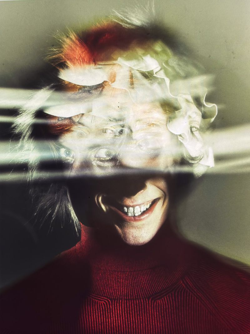 Abstract portrait of smiling adult with blonde, red, and green hair, wearing a red turtleneck sweater in a hazy, bright indoor setting.