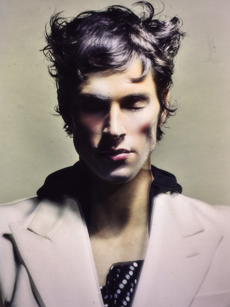 Portrait of a young adult male with dark hair, closed eyes, wearing a white jacket, against a light beige background in soft studio lighting.