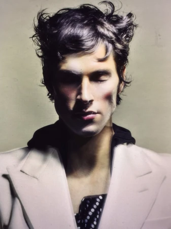Portrait of a young adult male with dark hair, closed eyes, wearing a white jacket, against a light beige background in soft studio lighting.