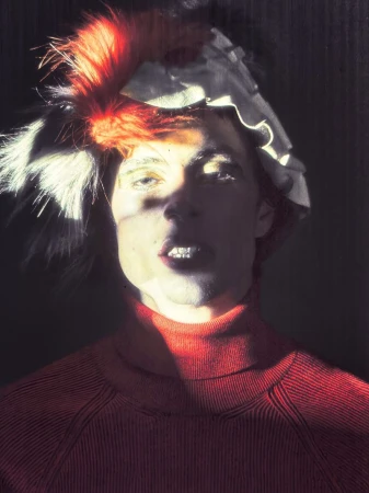 Portrait of an adult male wearing a red turtleneck sweater and a layered hat with white ruffles and red and white feathers.