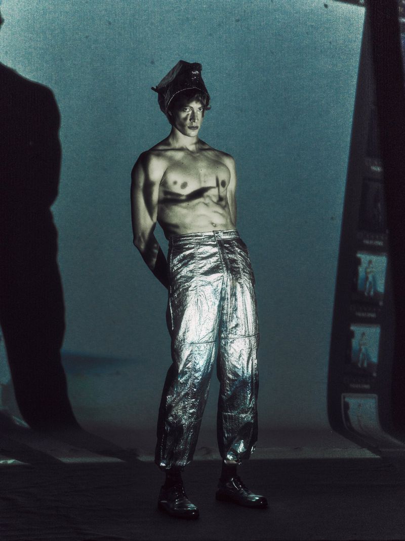 Shirtless male fashion model in silver metallic pants and head wrap poses in a moody, cinematic photo studio environment with shadows.
