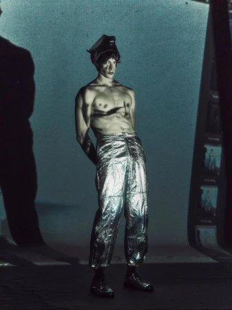 Shirtless male fashion model in silver metallic pants and head wrap poses in a moody, cinematic photo studio environment with shadows.