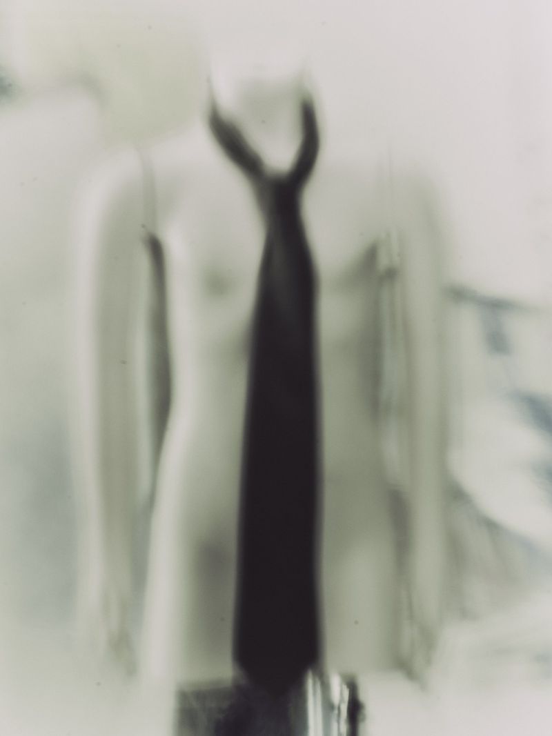 Blurry, desaturated photograph of a dark tie on a mannequin torso in front of a mottled, gray background with minimal detail.