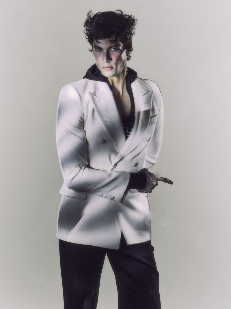 Fashion portrait of a stylish adult male in a white blazer, black hoodie, and gloves pointing to the right with a neutral expression.