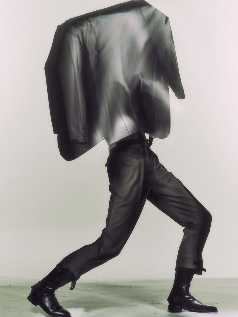 Surreal fashion image of a model walking in dark tailored trousers and black leather boots beneath an abstracted, motion-blurred blazer.