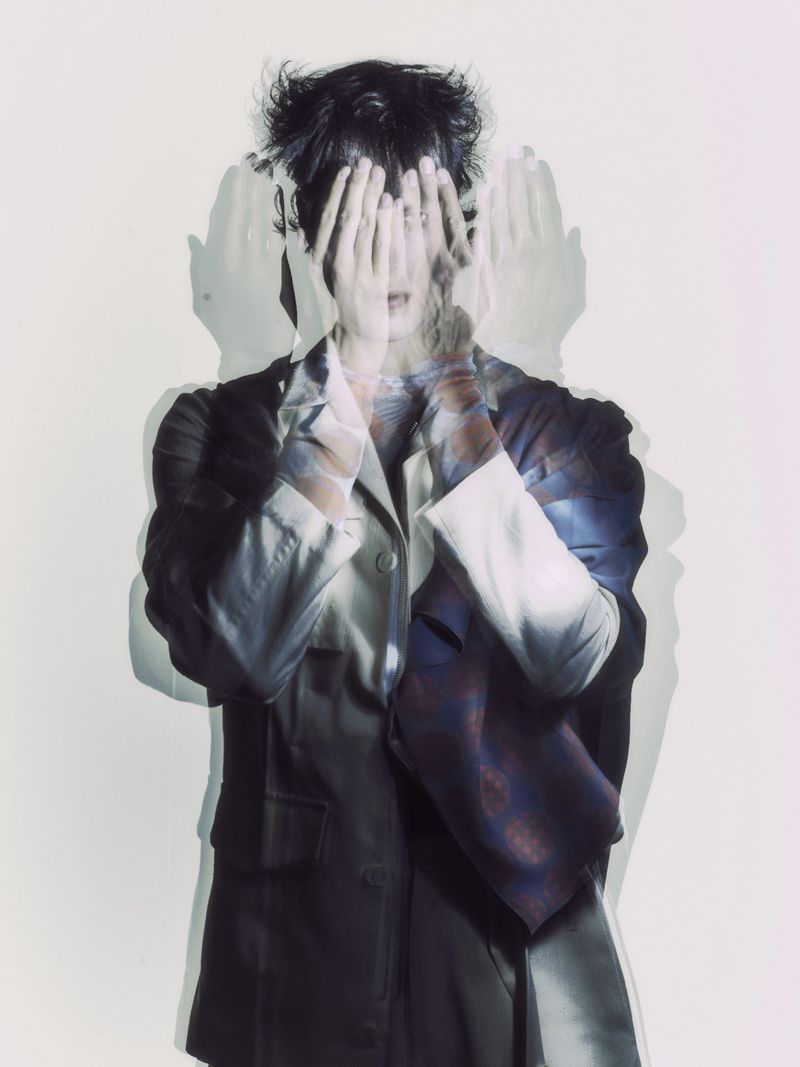 Multiple exposure portrait of a person covering their face with their hands, wearing a jacket, against a light background in a photo studio.
