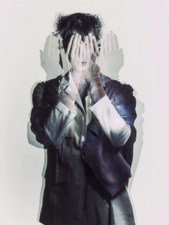 Multiple exposure portrait of a person covering their face with their hands, wearing a jacket, against a light background in a photo studio.